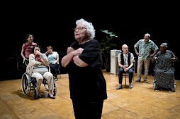 On center stage a woman is sharing a story and the rest of the cast is standing behind her.